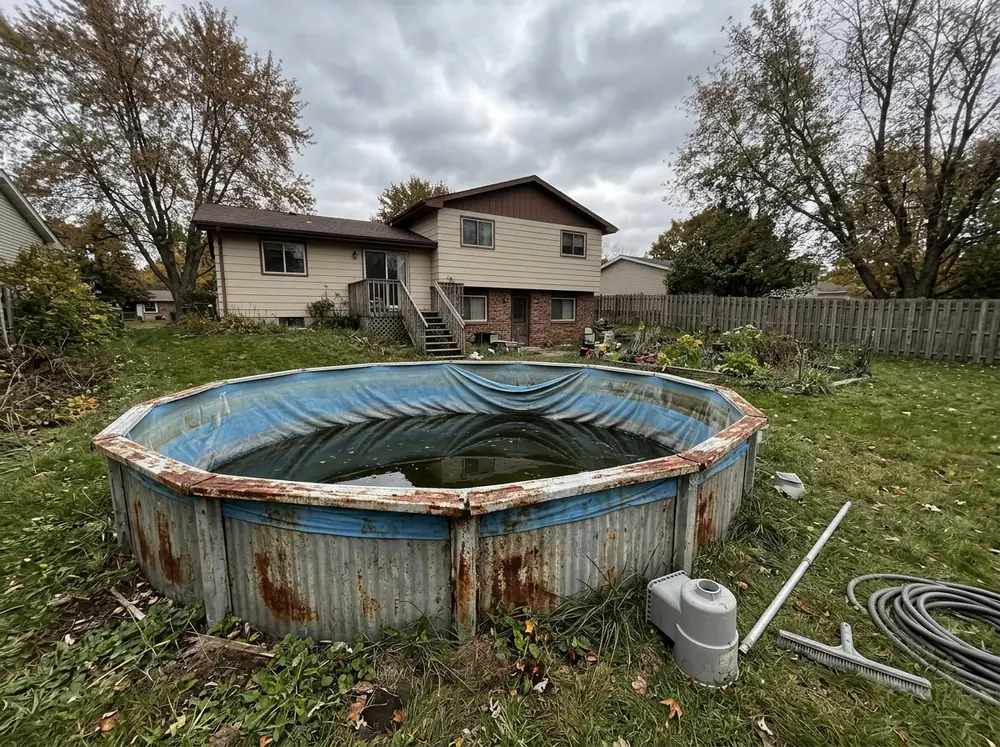 Round above-ground pool with a corrugated steel wall, rusted top rails and a sagging blue liner, half drained, in a backyard